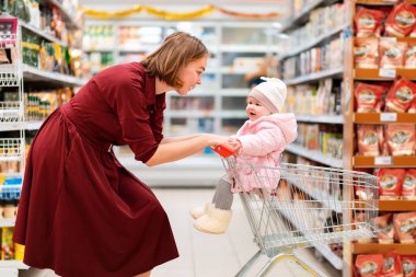 Genç bir annenin portresi marketin etrafında bir market arabası yuvarlıyor, içinde bebek oturuyor. Yan görüş. Aile alışverişi kavramı.