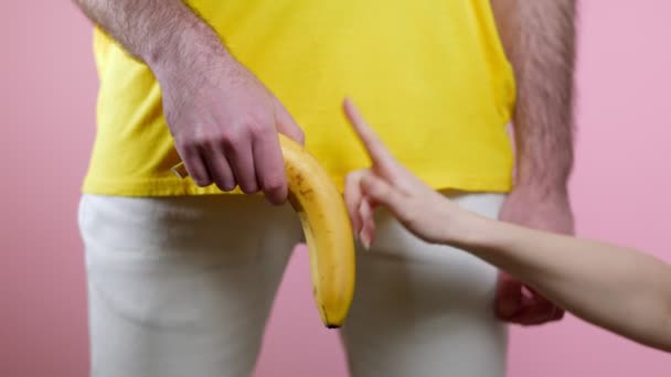 男は性器のレベルで彼の手に低いバナナを保持しています 女性の手はバナナの上に指を走らせ立ち上がる ピンクの背景 男性の健康の概念 — ストック動画