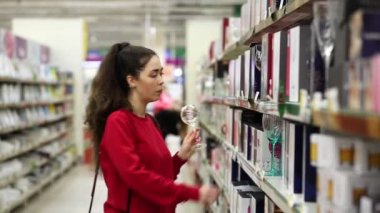 Genç ve güzel bir kadın mağazada şarap kadehlerini seçiyor ve karşılaştırıyor. Arka planda sıra sıra mallarla dolu bir koridor var. Yan görüş. Tüketim ve alışveriş kavramı