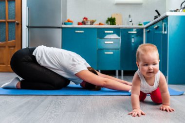 Evde yoga. Genç bir anne mindere yoga yapıyor. Bebeği paspasın üzerinde sürünüyor. Evde çocuklarla spor ve yoga.