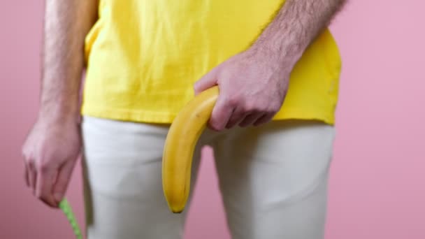 男は性器のレベルで彼の手に低いバナナを保持し 錠剤でそれをストロークし バナナが立ち上がる 閉じます ピンクの背景 男性の健康の概念 — ストック動画