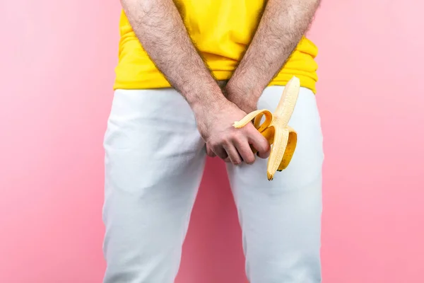 Potency and men\'s health. A man in white jeans, legs spread, covers his genitals with one hand, and holds an open banana with the other. Pink background. Close up.