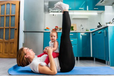 Bebekli bir anne mutfakta paspasın üstünde spor yapıyor, bacaklarını kaldırıyor. Yan görüş. Çocuklarla evde spor eğitimi kavramı.