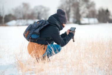 Sırtında sırt çantası olan genç bir kadın, çömelmiş ve telefonunda kurumuş çimen resimleri çekiyor. Yan görünüm.