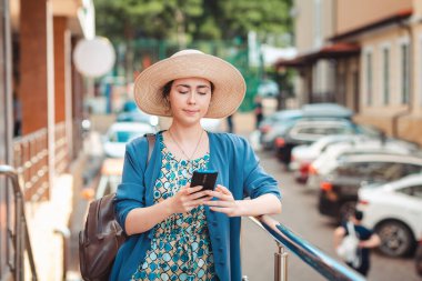 Şapkalı zarif bir kadın akıllı telefonunda bir şey yazıyor. Arka planda park yeri olan bir cadde var. İletişim ve sosyal ağlar kavramı.