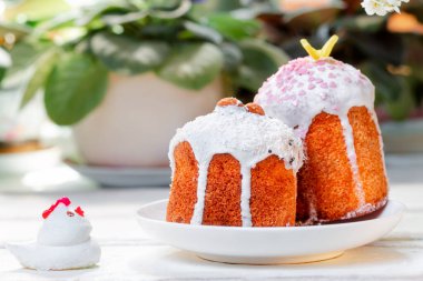 Tavuk heykelciğinin yanındaki bir tabakta iki süslü paskalya pastası. Arka planda bitkiler ve çiçekler var. Paskalya Bayramı.