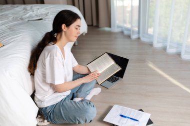 Çevrimiçi kurslar ve online öğrenme kavramı. Genç bir kadın yerde oturmuş kitap okuyor. Yanında bir laptop ve kağıtlar var..