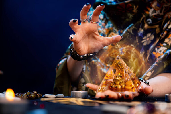 Предсказательница держит в руках стеклянную пирамиду с золотом и накладывает на неё заклятие. Волшебная аура. Крупный план. Принято. Понятие гадания, магии и эзотерики.