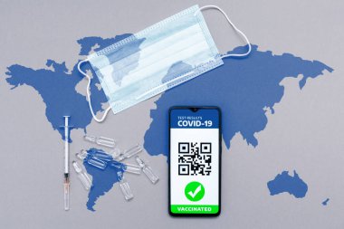 Aşı kavramı. Elektronik dokunulmazlık pasaportu, üzerinde COVID-19 aşı damgası olan akıllı telefon ekranında aşılar, koruyucu maske ve dünya haritasında şırınga. Düz yatıyordu.