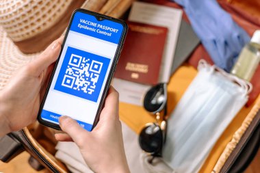 Bir kadının ellerine yakından bak. Bağışıklık sistemi dijital pasaportlu bir akıllı telefon tut. Arka planda, elbiseli bir bavul koruyucu maskeler ve eldivenler, bulanık bir hasır şapka. Düz yatıyordu.