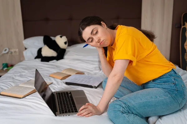 Concept of online courses and online education. Portrait of a young Caucasian woman is sitting with a bored look on the bed, and a laptop and books are lying next to her.