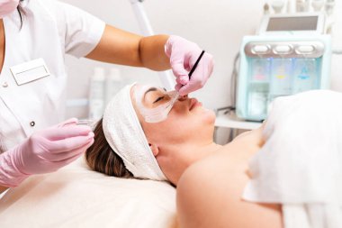 Lastik eldiven takan bir kozmetolog, müşterinin yüzüne krem maske takmak için fırça kullanır. Yakın plan. Arka planda bir kozmetoloji cihazı var. Kozmetoloji ve tedavi kavramı.