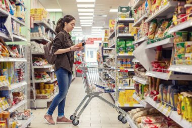 Genç ve güzel bir kadın market arabasını yuvarlar ve süpermarketten ürünler seçer..
