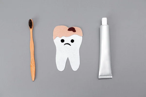 Toothpaste, a bamboo toothbrush, and a caries-ridden tooth carved out of felt with a cartoon face. Flat lay. Gray background. The concept of eco-friendly personal care products.