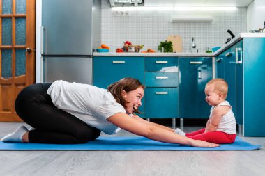 Evde yoga. Genç bir anne mindere asana yapıp bebeğinin üzerine yaslanıyor. Evde çocuklarla spor ve yoga..