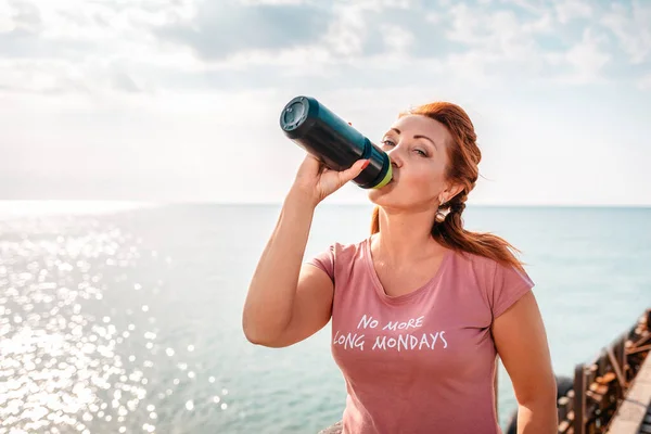 Portrait of an adult woman drinks from a shaker, posing against the background of the ocean and sky. Outdoor. Healthy lifestyle concept.