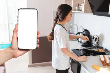 Genç bir kadın mutfakta kahvaltı hazırlıyor. Beyaz ekranlı akıllı telefonu tutan bir kadın eli. Model yap. Ev yapımı yemek ve yemek teslimatı..