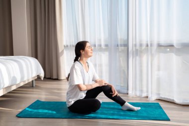 Yoga. Genç ve güzel bir kadın spor minderinde oturuyor, pencereden dışarı bakıyor ve sabah güneşine gülümsüyor. Yan görüş. Uzayı kopyala Ev egzersizi konsepti.