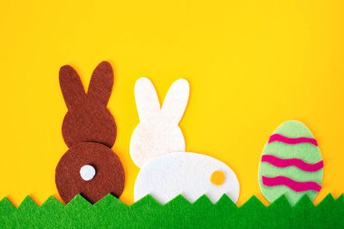 Paskalya yumurtası ve çimenlerin üzerinde beyaz ve kahverengi tavşanların keçeli uygulamalarından kesilmiş. Sarı arka plan. Dikiz aynası. Düz yatıyordu. Paskalya konsepti. Boşluğu kopyala.