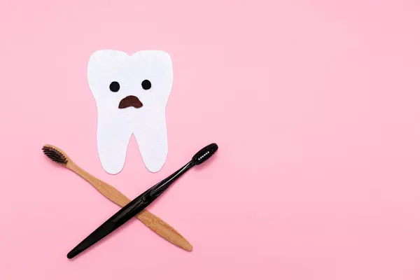 Care for the oral cavity. A startled tooth, cut out of felt with crossed toothbrushes, in the shape of a skull and bones. Flat lay. Pink background. Copy space. The concept of dental diseases.