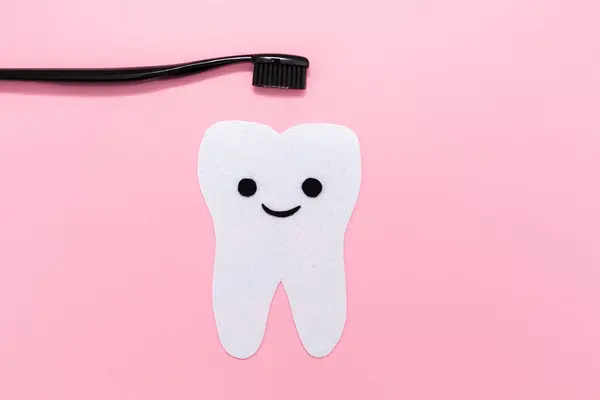 The tooth is carved and felt with a smiling cartoon face. Black plastic brush. Pink background. Flat lay. Copy space. Concept of oral hygiene.