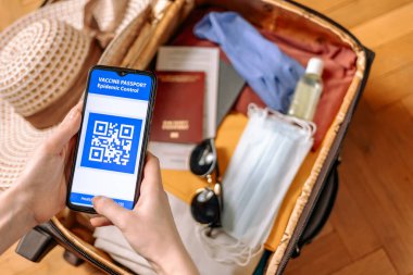 Bir kadının ellerinin en üst görüntüsü dijital pasaportlu bir akıllı telefonu tutuyor. Arka planda, elbiseli bir bavul koruyucu maskeler ve eldivenler, hasır şapka ve bulanık güneş gözlüğü. Yeni normal kavramı.
