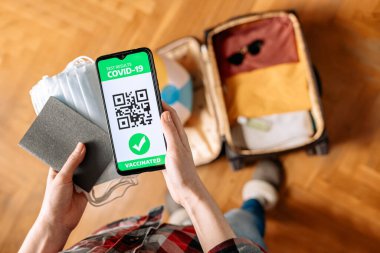 Yeni normal kavram. Bir kadının elinde pasaport, koruyucu maskeler ve bağışıklık sistemi dijital sağlık pasaportu olan bir akıllı telefon var. Arka planda, bulanık elbiseli bir bavul. Üst görünüm.