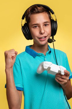 Sanal ve bilgisayar oyunları konsepti. Mavi tişörtlü ve kulaklıklı elinde joystickle internet oyunları oynayan genç bir çocuk. Zafer duygusu. Sarı arkaplan.