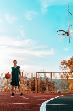 Spor ve basketbol. Mavi eşofmanlı bir genç bir top atmayı hedefleyen bir spor sahasında dikilirken topla poz veriyor. Arka planda mavi gökyüzü. Dikey. Boşluğu kopyala.