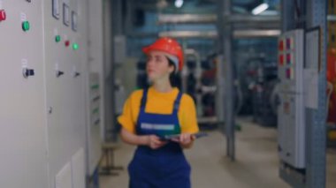 Koruyucu kask takan, elinde tabletle kameraya doğru yürüyüp gülümseyen genç bir kadın mühendis. Modern endüstri ve üretim