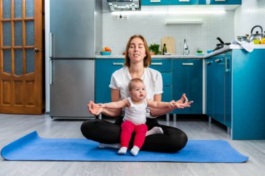 Genç bir anne mutfakta meditasyon yapıyor, spor minderinde oturuyor. Bebek annesinin ayaklarının üzerinde oturuyor. Yoga ve meditasyon kavramı.