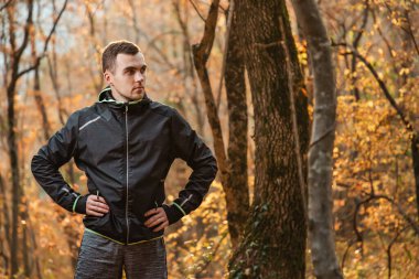 Spor anlayışı ve aktif yaşam tarzı. Sonbahar ormanında poz veren siyah eşofmanlı genç bir adam. Boşluğu kopyala.