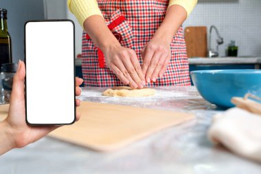 Önlüklü bir kadın mutfakta hamur topluyor. Solda bir kadının eli akıllı telefonu yakın planda tutuyor. Model yap. Kapatın. Ev yapımı gıda ve modern teknoloji kavramı..