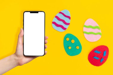 Paskalya konsepti. Renkli yumurtaların keçeli kumaşlarını kes. El ele tutuşarak cep telefonu. Model yap. Sarı arka plan. Boşluğu kopyala.