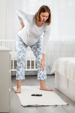 Hamile bir kadın sırt ağrısı ve yerde uzanmış bir akıllı telefonu kaldıracak kadar büyük bir mideyle zar zor eğilebilir. Hamile kadınların her gün yaşadığı zorluklar. Dikey.