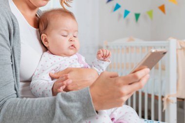 Bir anne uyuyan bir çocuğu kollarında tutar ve akıllı telefonuna yazar. Bebeğin yakın plan portresi. Çevrimiçi bağımlılık kavramı.