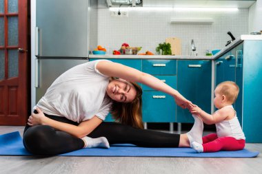 Genç bir anne ve bebeği mutfakta bir paspasın üzerinde esniyorlar. Çocuk annenin bacağının yanında oturuyor. Çocuklarla evde spor eğitimi kavramı.