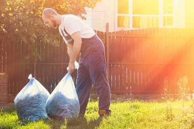 Mavi üniformalı bir adam gün batımında temizliği bitirir, gübre torbaları toplar. Güneş sağda parlar..
