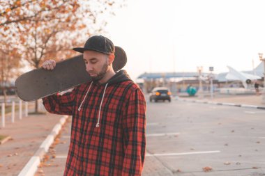 Kaykay yapmak. Sakallı, şapkalı, şık giyinmiş elinde kaykayla poz veren yakışıklı bir adam. Arka planda, şehrin boş bir caddesi. Boşluğu kopyala.
