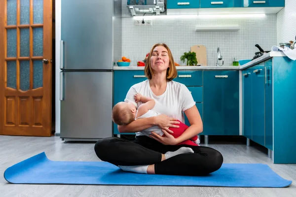 Yoga at home. A young smiling mother meditates in the kitchen, sitting on a sports mat, holding the baby in her arms. The concept of yoga and meditation with children.