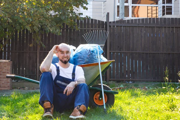 The gardener in blue uniform is resting on the grass. Smile and good mood. Garden supplies in the background.