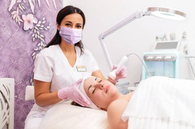 Güzellik salonu. Lastik eldiven takan bir kozmetolog kaldırma cihazı kullanarak gençleştirici bir prosedür uyguluyor. Arka planda kozmetik bir cihaz var. Profesyonel kozmetoloji ve güzellik kavramı.