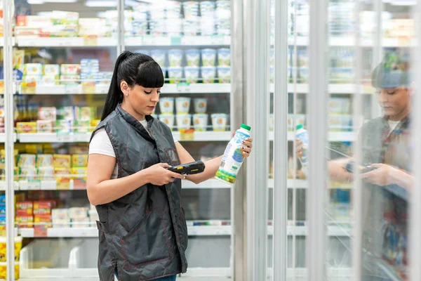 A young Caucasian woman working as a Manager in a supermarket checks the expiration date of milk in a bottle, using an electronic device. Business and technology concepts.
