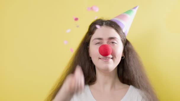 紙の帽子を被った陽気な女性と 喜んでコンフェッティを投げる偽の赤い鼻 黄色の背景 4月のバカの日のコンセプト — ストック動画