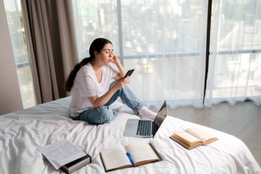 Genç, beyaz bir kadın bir yatakta oturuyor ve akıllı telefon kullanıyor. Laptop ve kitaplar yakında. Uzayı kopyala Çevrimiçi eğitim ve sosyal ağlara bağımlılık kavramı.