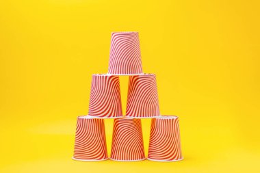 Sarı zemin üzerinde çizgili kırmızı kağıt bardaklardan oluşan bir piramit. Boşluğu kopyala.