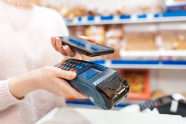 Bir kadının elleri ödeme terminalini tutuyor ve akıllı telefondan alışveriş için para ödüyor. Telefon ekranında, wi-fi ağında. NFC, iş ve bankacılık işlemleri kavramı.