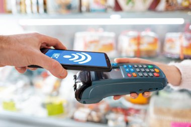 Satıcı ödeme terminalini elinde tutuyor ve satın alma parasını akıllı telefon kullanarak ödüyor. Yan görüş. Telefon ekran kablosuz ağıyla. NFC konsepti, iş ve bankacılık operasyonları.