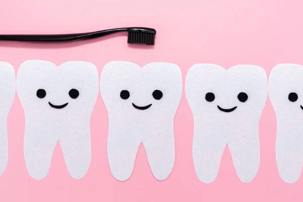 Concept of oral hygiene. The teeth cut out of felt with a smiling cartoon face. Black plastic brush. Pink background. Flat lay. Copy space.