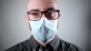 Gözlük takan beyaz bir adam ve yüzünde tıbbi maske olan siyah bir gömlek doğrudan kameraya bakıyor. Gri arka plan. Viral enfeksiyonlara ve soğuk algınlığına karşı korunma kavramı.